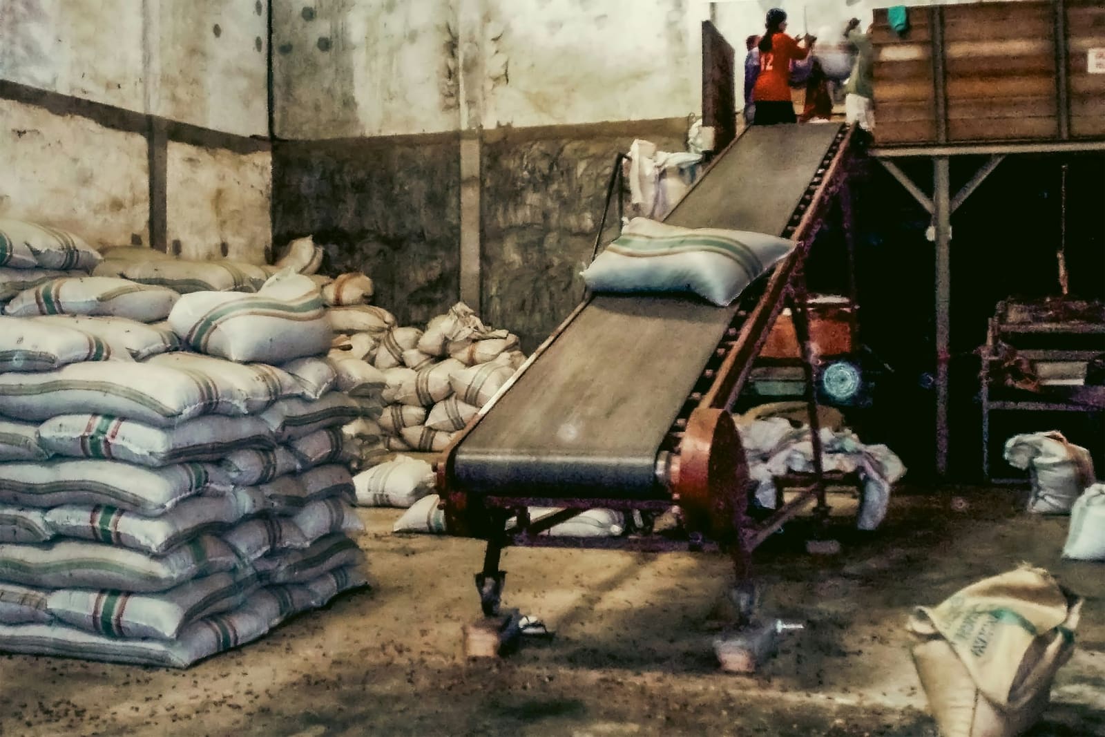 Sacks stacked in a mill warehouse beside a conveyor running up to a loading platform.
